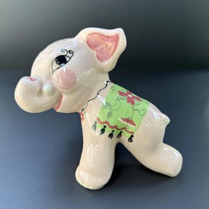 May include: A glossy, light pink ceramic elephant figurine featuring a painted green blanket on its back with red floral patterns and black fringe details. The elephant has large ears with pink accents, a curled trunk, and black painted eyes.