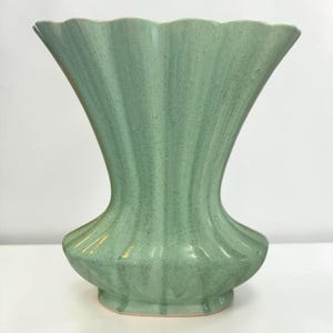 May include: A light green ceramic vase with a speckled finish and a fluted, scalloped rim. The vase features a wide, flared opening that tapers down to a narrower neck, expanding into a rounded base with vertical ridges throughout the body.