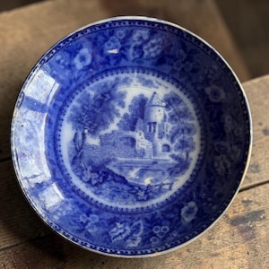 May include: A blue and white antique plate with a detailed landscape scene in the center. The plate features a castle, trees, and a bridge. Floral patterns adorn the rim. The plate is round and appears to be made of ceramic.