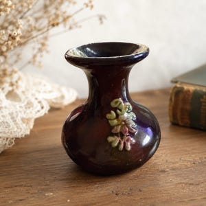 May include: A small, dark purple ceramic vase with a glossy finish and a wide, flared rim. The body of the vase features a raised, three-dimensional floral relief decoration in muted green and pink tones on the front surface.