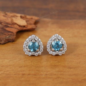Könnte beinhalten: Ein Paar Ohrstecker mit Tropfenform. Jeder Ohrring hat einen runden, hellblauen Edelstein, der von einem Kranz aus kleineren, klaren Edelsteinen umgeben ist. Die Ohrringe liegen auf einer Holzoberfläche.