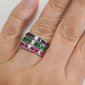 Może przedstawiać: Trzy nakładane na siebie pierścienie z rzędem kwadratowych kamieni szlachetnych. Górny pierścień ma niebieskie szafiry, środkowy pierścień ma zielone szmaragdy, a dolny pierścień ma różowe rubiny. Każdy pierścień ozdobiony jest małym diamentem.