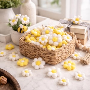 May include: A woven basket overflowing with white and yellow daisy-shaped decorations. Scattered around the basket are more of the same decorations. A small pot of daisies and a decorative bee are in the background. The daisies have white petals and yellow centers.