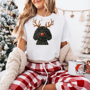 May include: White t-shirt with a black dog illustration wearing reindeer antlers and a red nose, a festive design for Christmas. The model is wearing red and white checked pyjama bottoms. The image has a Christmas theme.