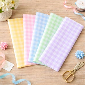 May include: Five sheets of pastel-colored gingham patterned paper in yellow, pink, blue, green, and purple. The sheets are fanned out on a wooden surface. Decorative elements include flowers, ribbons, and gold scissors.