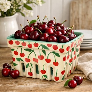 May include: A rectangular ceramic basket filled with ripe, dark red cherries. The basket is cream-colored with a cherry pattern in red and green. The basket sits on a wooden surface, with additional cherries scattered around.
