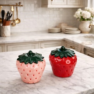 Puede incluir: Dos tarros de cerámica con forma de fresa y tapas verdes con hojas. Uno es rosa claro con puntos rojos, el otro es rojo brillante. Se encuentran sobre una encimera de mármol en una cocina.