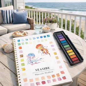 May include: A watercolor workbook titled "Seaside" with jellyfish illustrations and a color palette guide. A black watercolor paint set and a seashell are on a wooden table. The scene is set outdoors with a view of the ocean.