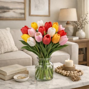 May include: A vibrant bouquet of tulips in a clear glass vase. The tulips display a variety of colors, including red, yellow, pink, and white. The vase sits on a marble-patterned table, with a candle and decorative beads nearby. The scene is set in a living room.