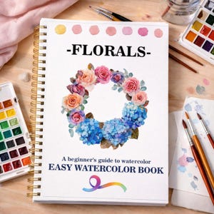 May include: A watercolour book titled "-FLORALS-" with a floral wreath design of roses and hydrangeas. The book includes the text "A beginner's guide to watercolour" and "EASY WATERCOLOR BOOK". Watercolour paints and brushes are visible.