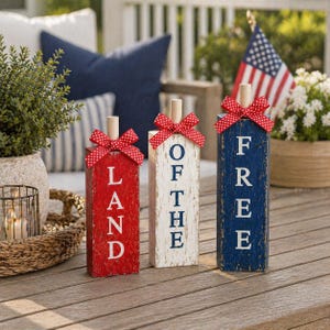 May include: Three wooden decorative blocks in red, white, and blue, with the words "LAND", "OF THE", and "FREE" painted vertically. Each block is topped with a wooden dowel and a red bow with white polka dots. The blocks are on a wooden table.