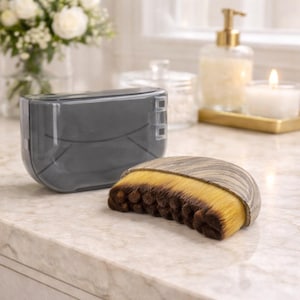 May include: A gray makeup brush cleaner with a matching case. The brush has a wooden-look handle and yellow and brown bristles. The case is rectangular with rounded edges and a clear lid.