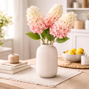 May include: A white, ribbed ceramic vase holds three large hydrangea stems with pink and white ombre blooms and green leaves. The vase sits on a wooden table with a neutral-colored runner, with books and a ball of twine nearby.