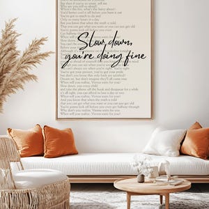 May include: A framed print with the words "Slow down, you're doing fine" in a handwritten font. The print hangs above a white sofa with orange and white cushions. A round wooden coffee table and woven chair are in front.