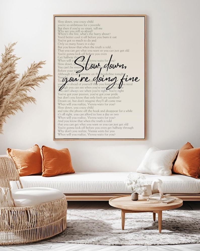 May include: A framed print with the words "Slow down, you're doing fine" in a handwritten font. The print hangs above a white sofa with orange and white cushions. A round wooden coffee table and woven chair are in front.