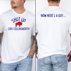 May include: White t-shirt with blue text that reads "SHUT UP" above a red bison graphic and the name "CRIS COLLINSWORTH". The back of the shirt has the text "NOW HERE'S A GUY...." in blue. The shirt is a casual style.