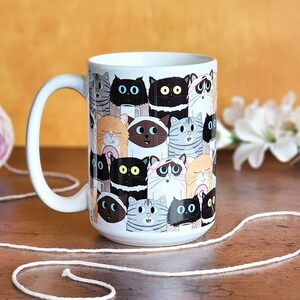 May include: White ceramic mug with a colorful pattern of cartoon cats. The cats are all different colors and have different expressions.