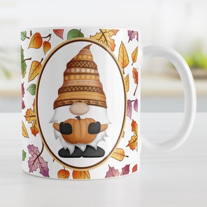 May include: White ceramic coffee mug with a fall-themed design. The mug features a gnome holding a pumpkin and surrounded by colorful autumn leaves.