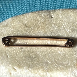 May include: A gold-toned metal safety pin brooch with a smooth, elongated rectangular frame. The pin features a classic coil mechanism at one end and a secure clasp at the other, with a small, stamped hallmark visible on the central bar.