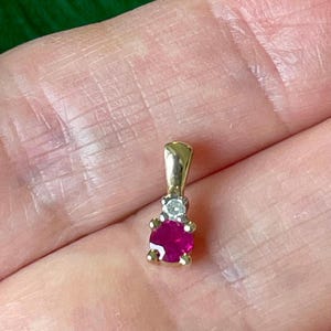 May include: A gold pendant featuring a round, vibrant pink gemstone set below a small, clear, round-cut diamond. The pendant has a polished gold bail at the top, designed for attachment to a chain, showcasing a classic two-stone vertical arrangement.