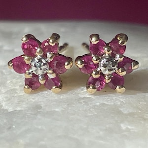 May include: A pair of flower-shaped stud earrings. Each earring features a cluster of pink gemstones forming petals around a central, sparkling stone. The earring settings are gold-colored.