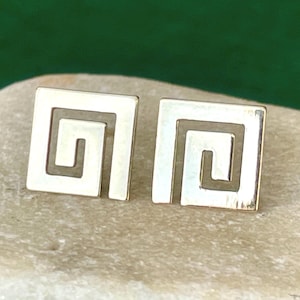 May include: A pair of gold-toned stud earrings, each featuring a square shape with a cut-out Greek key or meander pattern. The earrings have a polished, reflective metallic finish and are presented side-by-side, showcasing the geometric design.