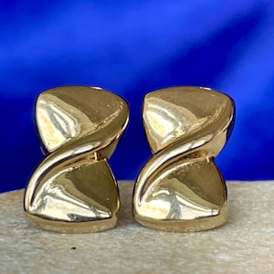 May include: A pair of polished gold-tone stud earrings featuring a smooth, abstract hourglass shape with a diagonal band crossing the centre. The metal surface is highly reflective, creating bright highlights and deep shadows across the curved form.