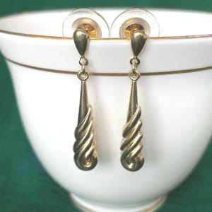 May include: A pair of gold drop earrings featuring a smooth teardrop-shaped top section connected to a textured, spiraling, fan-like base. The earrings include clear plastic earring backs and exhibit a polished, reflective metallic finish throughout.