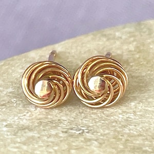 May include: A pair of gold-colored stud earrings. Each earring features a central, rounded stud surrounded by a spiral design. The earrings are displayed on a light-colored surface.