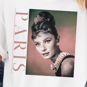 Può includere: Maglietta bianca con una stampa grafica di Audrey Hepburn in un abito nero con una collana di perle. Il testo "PARIS" è stampato verticalmente sul lato sinistro dell'immagine.