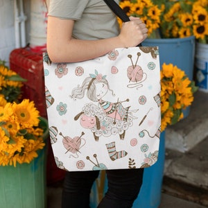 May include: A white tote bag with a colorful pattern of a girl knitting on a sheep. The pattern includes yarn balls, knitting needles, and flowers. The bag has black straps.