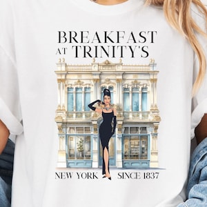 Könnte beinhalten: Weißes T-Shirt mit einem Grafikdruck eines Gebäudes mit dem Text "Breakfast at Trinity's, New York, Since 1837". Eine Frau in einem schwarzen Kleid steht vor dem Gebäude.