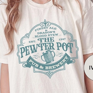 May include: A white t-shirt with a vintage-style graphic of a pub called "The Pewter Pot" featuring a mug of ale and the text "Finest Ale & Dragon's Blood Stew Est. 1347 Pub & Brewery". The shirt is labelled "Ivory".