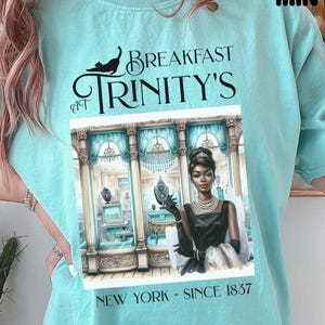 Könnte beinhalten: Ein türkisfarbenes T-Shirt mit einem Grafikdruck einer Frau in einem schwarzen Kleid, die vor einem Schaufenster steht. Der Text auf dem Shirt lautet "Breakfast at Trinity's, New York - Since 1837".