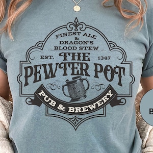 May include: A blue t-shirt featuring a vintage-style logo for "The Pewter Pot Pub & Brewery". The logo includes a pewter tankard and the text "Finest Ale & Dragon's Blood Stew, Est. 1347".