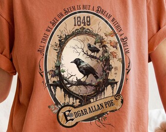May include: A burnt orange t-shirt with a vintage-style graphic featuring two black birds perched on a branch. The graphic includes the text "All that we see or seem is but a dream within a dream" and "1849" with the name "Edgar Allan Poe" at the bottom.