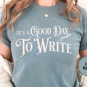 May include: A light blue t-shirt with the text "It's a Good Day To Write" in white lettering. The text is styled with a decorative flourish and a feather. The t-shirt is being worn by a person.