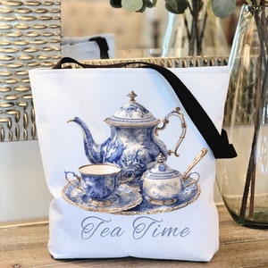 Puede incluir: Bolsa de tela blanca con asa negra. La bolsa presenta una tetera, un azucarero y una taza de té azules y blancos con ribete dorado. El texto "Tea Time" está impreso en negro en la parte inferior de la bolsa.