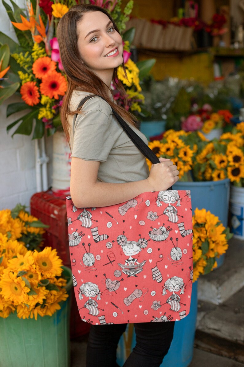 May include: Red tote bag with a repeating pattern of knitting-themed illustrations. The design includes cartoon figures, yarn balls, knitting needles, and socks. The bag has a black strap and is being held in front of a flower shop.