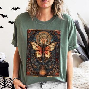 May include: A green t-shirt with a colorful butterfly graphic. The butterfly is surrounded by a stained glass-like design with intricate details. The design is reminiscent of Art Nouveau style.