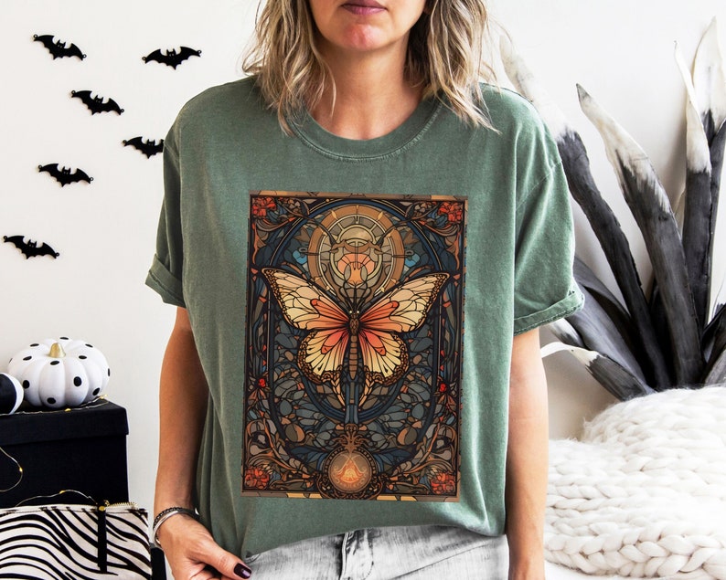 May include: A green t-shirt with a colorful butterfly graphic. The butterfly is surrounded by a stained glass-like design with intricate details. The design is reminiscent of Art Nouveau style.