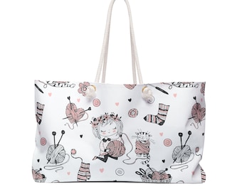 May include: White canvas tote bag with a repeating pattern of a girl knitting, a cat, yarn balls, knitting needles, and hearts. The bag has white rope handles.