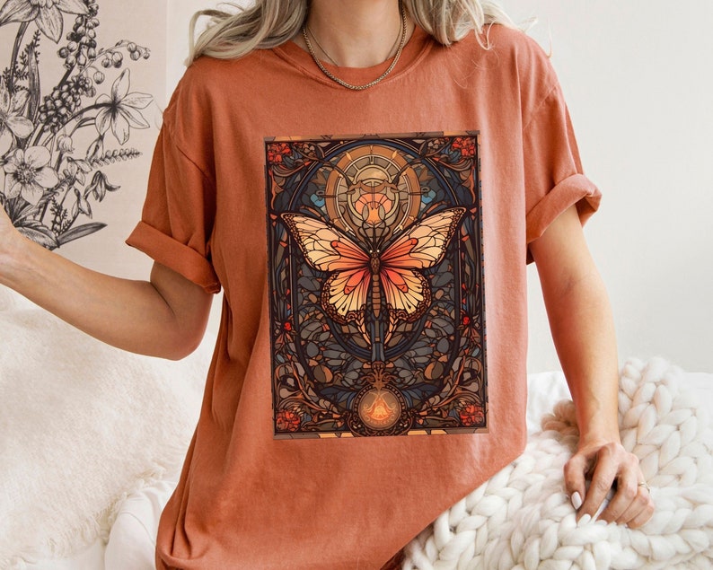 May include: A rust-colored t-shirt with a graphic print of a butterfly in a stained glass style. The butterfly has orange, yellow, and brown wings. The background is a dark blue and brown stained glass design.