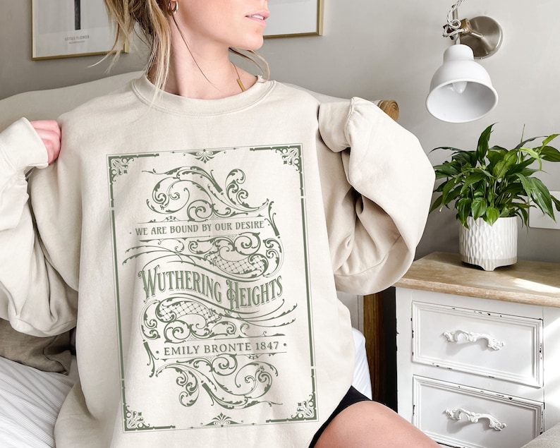 May include: A beige sweatshirt with a vintage-style graphic print of the words "Wuthering Heights" with decorative flourishes. The text also includes "We are bound by our desire" and "Emily Bronte 1847".