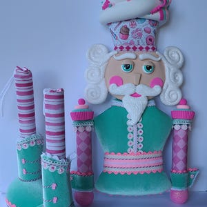 May include: A whimsical nutcracker figurine with a pink and green candy-themed design. The nutcracker has a white cupcake hat with pink and green frosting and sprinkles, a white beard, and a green velvet body with pink and white trim. The nutcracker is holding two pink and white candy cane legs.