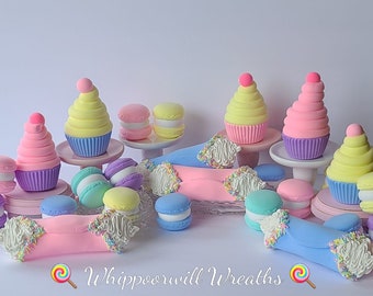 May include: A collection of pastel-colored cupcakes and macarons, decorated with sprinkles and frosting. The cupcakes are topped with a swirl of frosting and a pink or yellow ball. The macarons are in shades of blue, pink, and yellow. The image includes the text "Whipporwill Wreaths".