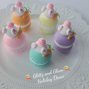 May include: Five pastel-colored macarons decorated with fondant bunnies and carrots. The macarons are arranged on a white plate with a decorative border. The text "Glitz and Glam Holiday Decor" is visible in the bottom right corner of the image.