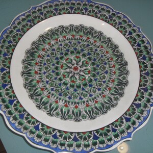 May include: A white ceramic plate with a scalloped edge and a colourful floral design. The design features green, blue, red, and white flowers and leaves.