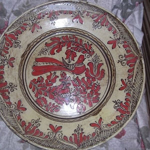 Puede incluir: Un plato de cerámica blanco con un diseño floral rojo y marrón. El plato presenta un pájaro estilizado con una cola larga y un patrón floral alrededor del borde.