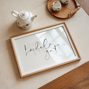 Könnte beinhalten: Ein gerahmter Druck mit dem Text "Dein Lächeln steht dir gut" in schwarzer Schrift auf weißem Hintergrund. Der Druck ist in einem hellen Holzrahmen und steht auf einem Tisch mit einer Teekanne und zwei Teetassen.